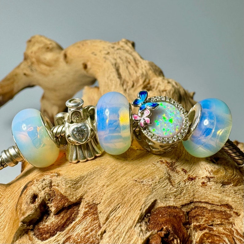 May include: A silver charm bracelet with five charms. The charms are a white opal bead, a silver angel charm with a clear crystal heart, a white opal bead, a silver charm with a butterfly and a pink flower, and a white opal bead.