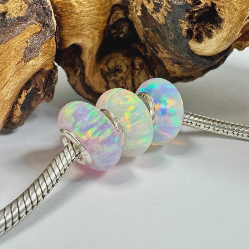 May include: Three iridescent opal beads with silver accents are strung on a silver chain. The opals display a range of colors, including pink, blue, and green. The background features a piece of wood.