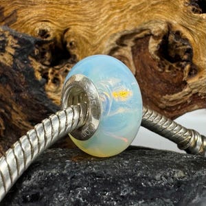 May include: A white opal bead with a silver charm bracelet. The bead has a light blue and yellow iridescent sheen. The charm bracelet is silver and has a twisted design.
