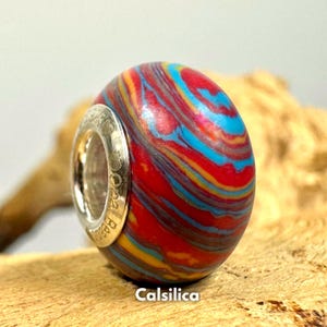Puede incluir: Una cuenta con un patrón rojo, azul y amarillo con un dije de plata. El dije tiene la palabra "Calsilca" grabada.