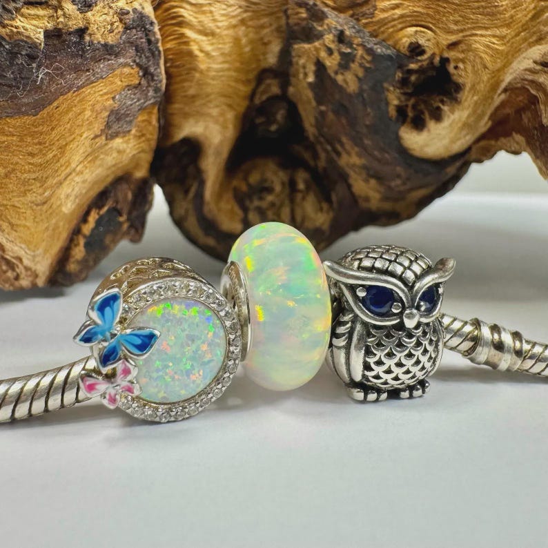 May include: A collection of silver charm beads. One bead features a butterfly design with blue and pink accents. Another is an opal-like bead. The third is a silver owl charm with blue eyes.