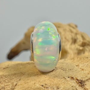 May include: A white opal bead with iridescent flashes of green, blue, and pink. The bead is encased in a silver-colored metal setting. The opal is the focal point, showcasing its natural colors and textures. The bead is a jewelry component.