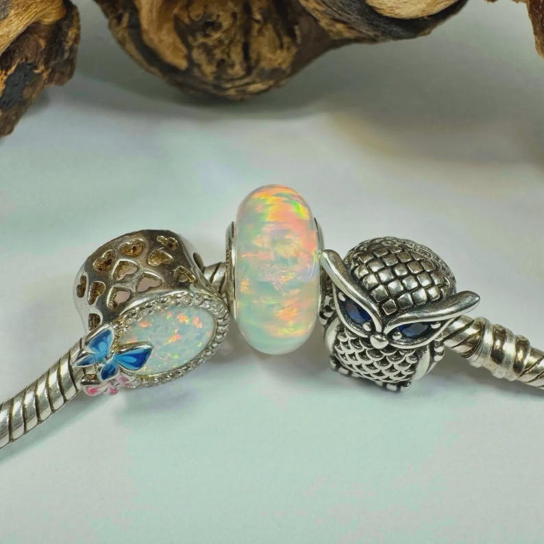 May include: A collection of silver charm beads. One bead is a white opal with rainbow hues. Another is a silver owl with blue eyes. The third charm has a butterfly and opal design.