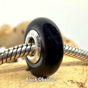 May include: A black obsidian bead with a silver core, on a silver chain bracelet. The bead is round and has a smooth, polished surface. The bead is a popular choice for jewellery making and is believed to have protective properties.
