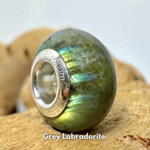 Puede incluir: Una cuenta de labradorita gris con reflejos verdes y azules iridiscentes. La cuenta está engastada en un engaste de plata con la palabra "Pandora" grabada en el lateral.