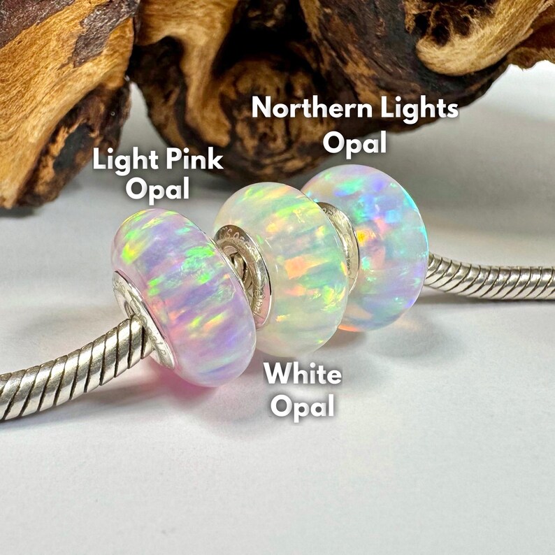 May include: Three opal beads in light pink, white, and Northern Lights colors are displayed on a silver chain. The beads have a shimmering, iridescent quality. Text on the image identifies each bead's color.