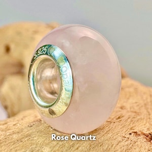 Puede incluir: Una cuenta de cuarzo rosa de color rosa claro con una banda plateada alrededor del centro. La banda está grabada con "Opal Beads" y "5.925".