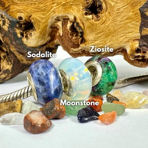 May include: Four gemstone beads on a silver bracelet, including sodalite, moonstone, ziosite, and a white bead. The beads are surrounded by small, colorful gemstones.