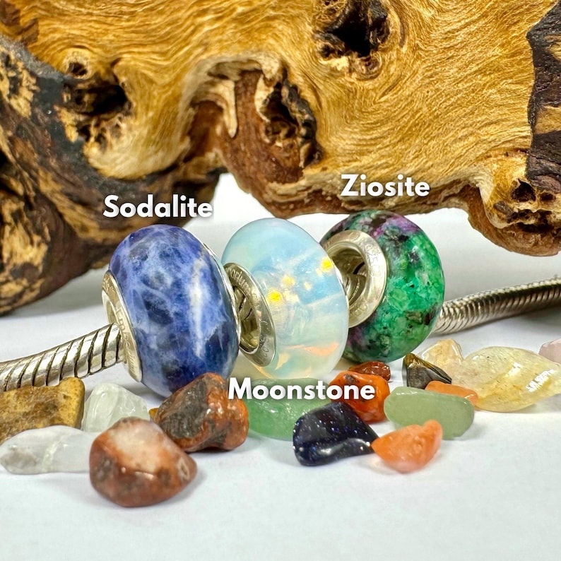 May include: Four gemstone beads on a silver bracelet, including sodalite, moonstone, ziosite, and a white bead. The beads are surrounded by small, colorful gemstones.