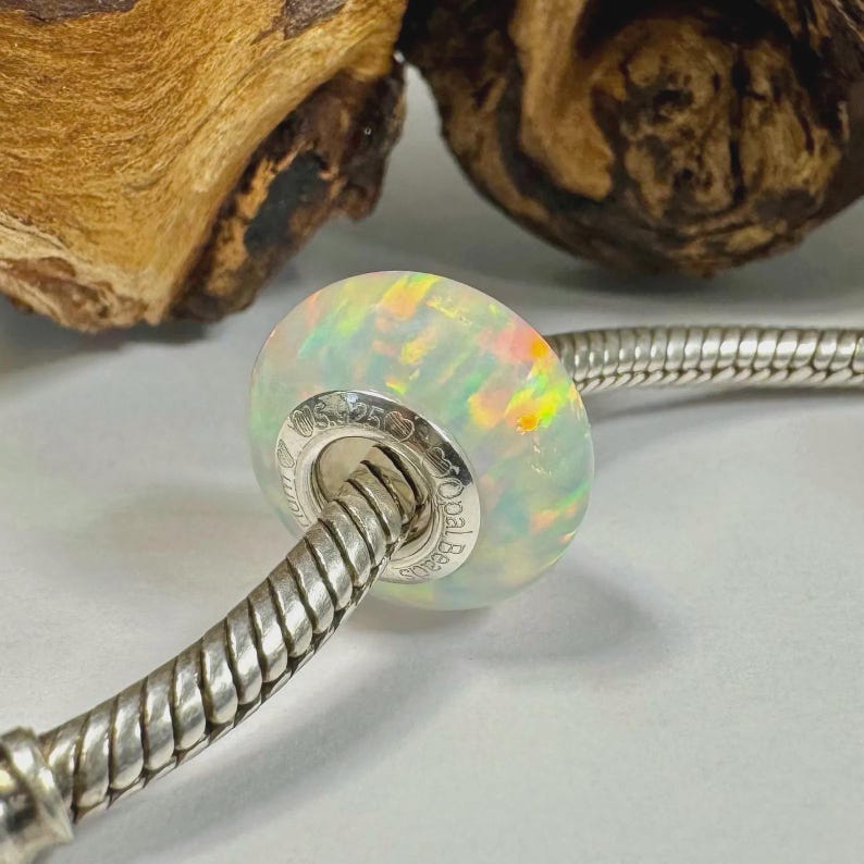 May include: A round, iridescent opal bead with a silver core is strung on a silver bracelet. The opal bead has a pearly white base color with flashes of yellow, orange, and green. The bead is marked with the text "S925" and "Opal Beauty".