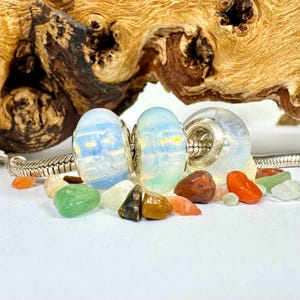 May include: Two iridescent white glass beads with silver accents and a silver charm bead. The beads are on a silver chain bracelet and surrounded by small colorful stones.