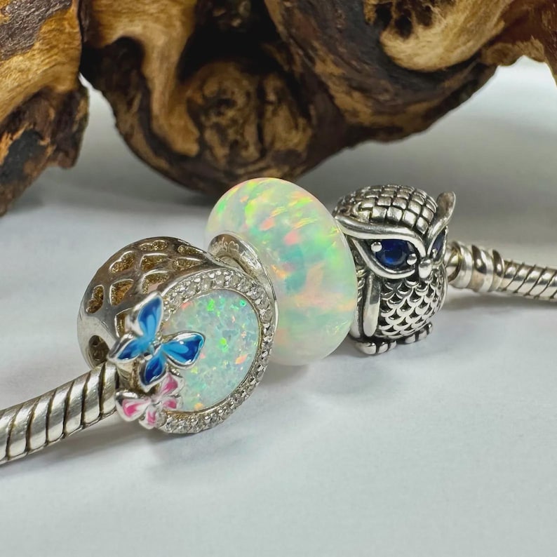 May include: A silver charm bracelet with three decorative charms. The charms include a butterfly charm with blue and pink enamel, an opal bead, and a detailed owl charm with blue eyes. The charms are on a silver bracelet.