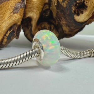 May include: A white opal bead with iridescent flashes of color is strung on a silver bracelet. The bead has a smooth, rounded shape and is the focal point of the image. The bracelet is made of a woven silver chain.