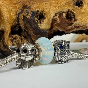 May include: Three silver charm beads for a bracelet. The bead on the left is a detailed silver charm of a character from Star Wars. The middle bead is a clear glass bead with an iridescent finish. The bead on the right is a silver owl charm with a blue gemstone eye.