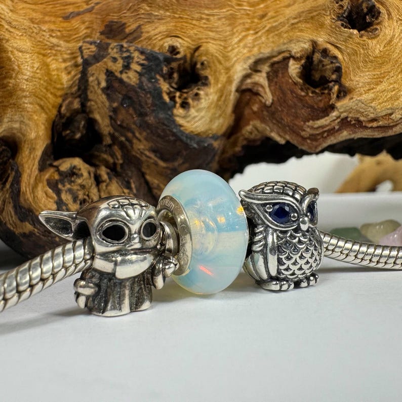 May include: Three silver charm beads for a bracelet. The bead on the left is a detailed silver charm of a character from Star Wars. The middle bead is a clear glass bead with an iridescent finish. The bead on the right is a silver owl charm with a blue gemstone eye.