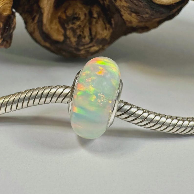 May include: A white opal bead with iridescent flashes of color, set on a silver-toned bracelet. The bead has a rounded shape and a polished surface, showcasing a play of light. The bracelet has a textured, snake-like design.