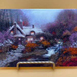 May include: A cottage with a thatched roof and lit windows sits in a snowy forest with a stream flowing in front of it. The scene is painted in a soft, dreamy style with purple, orange, and green hues.