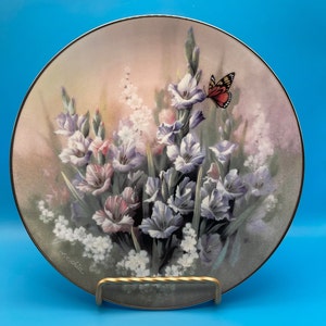 May include: A decorative ceramic plate with a floral design featuring a bouquet of purple and pink gladiolus flowers with a single orange butterfly. The plate has a gold rim and is signed "F.C. Schmidt".