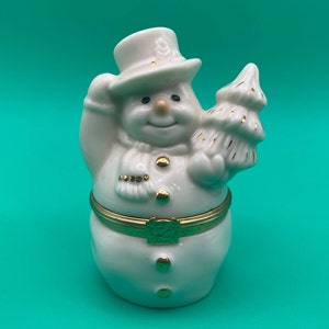 May include: A white porcelain snowman figurine with a gold hat, scarf, and buttons. The snowman is holding a gold-trimmed Christmas tree.
