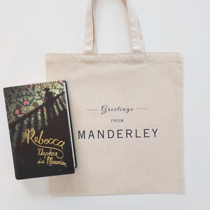 Beige canvas tote bag with the text Greetings from Manderley in black font. The tote is displayed flat on a white background with the book Rebecca by Daphne du Maurier placed beside it.