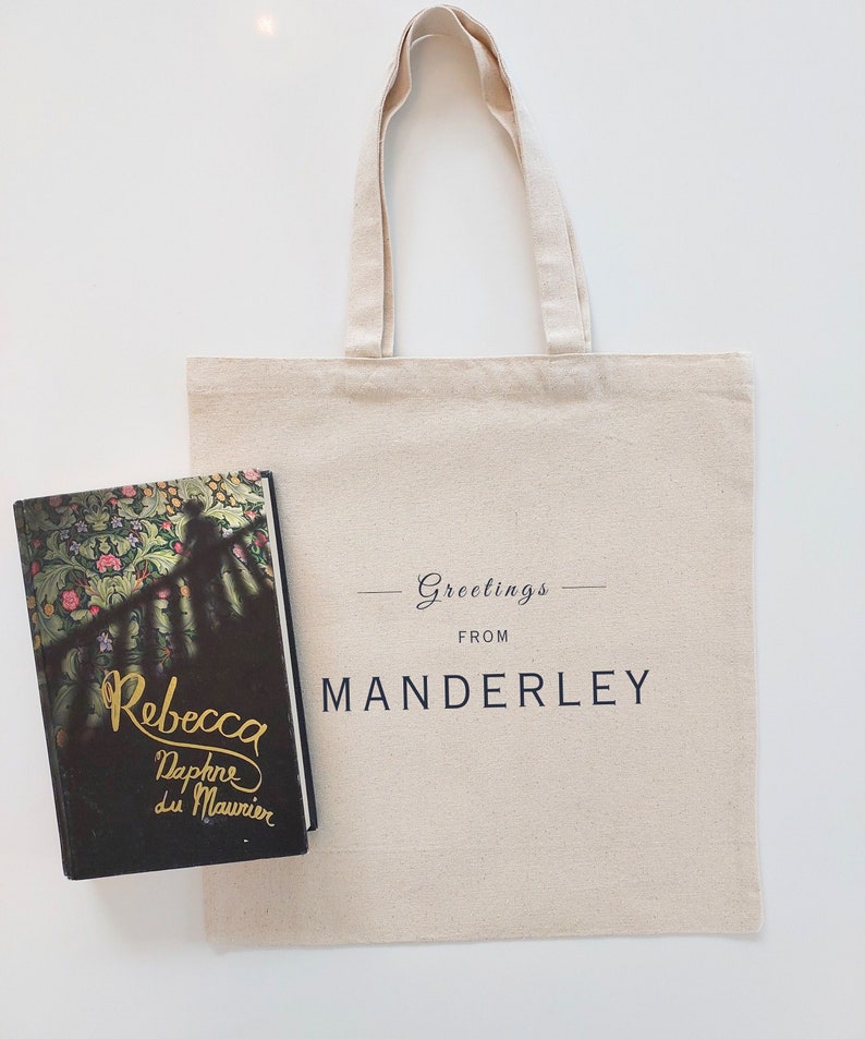 Beige canvas tote bag with the text Greetings from Manderley in black font. The tote is displayed flat on a white background with the book Rebecca by Daphne du Maurier placed beside it.