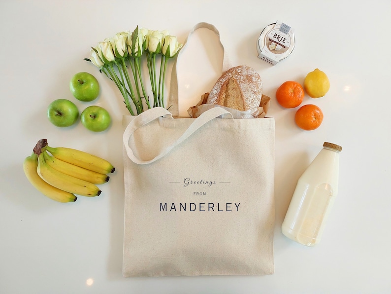 Beige canvas tote bag with the text Greetings from Manderley in black font.  It is displayed on a white background as a grocery bag with flowers, a loaf of bread, apples, bananas, milk, cheese, oranges and a lemon beside it.