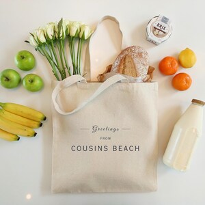 May include: A canvas tote bag with the text "Greetings from Cousins Beach" is filled with groceries. The bag contains a loaf of bread, white roses, and a bunch of bananas. Green apples, oranges, a lemon, a bottle of milk, and a wheel of brie cheese are also present.