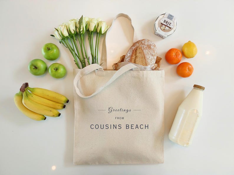 May include: A canvas tote bag with the text "Greetings from Cousins Beach" is filled with groceries. The bag contains a loaf of bread, white roses, and a bunch of bananas. Green apples, oranges, a lemon, a bottle of milk, and a wheel of brie cheese are also present.