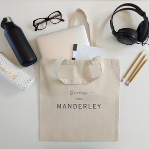 Beige canvas tote bag with the text Greetings from Manderley in black font. The bag is displayed on a white background and used as a college or work tote with a laptop, notebook, water bottle, headphones, glasses, pencil case and pencils.