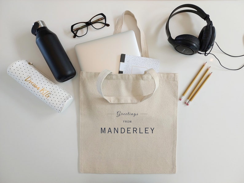 Beige canvas tote bag with the text Greetings from Manderley in black font. The bag is displayed on a white background and used as a college or work tote with a laptop, notebook, water bottle, headphones, glasses, pencil case and pencils.
