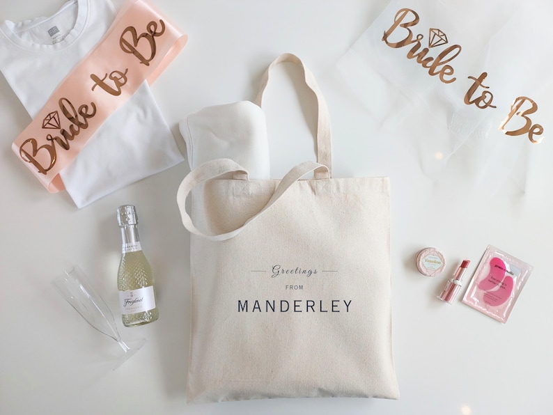 May include: A white tote bag with the text "Greetings from Manderley" printed on it. The bag is filled with a variety of items, including a white t-shirt, a pink sash that says "Bride to Be", a bottle of champagne, and a pair of eye masks.
