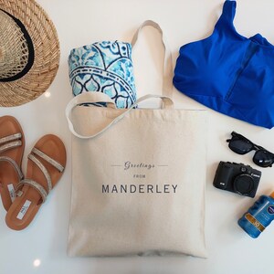 Beige canvas tote bag with the text Greetings from Manderley in black font. The bag is displayed on a white background and used as a beach bag with a straw hat, sandals, bikini top, beach towel, sunglasses, camera and sunscreen next to it.