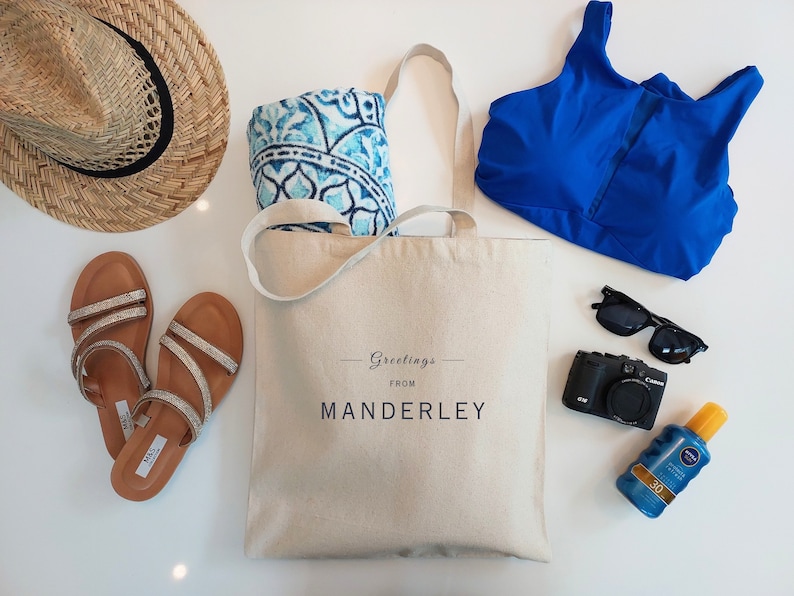 Beige canvas tote bag with the text Greetings from Manderley in black font. The bag is displayed on a white background and used as a beach bag with a straw hat, sandals, bikini top, beach towel, sunglasses, camera and sunscreen next to it.