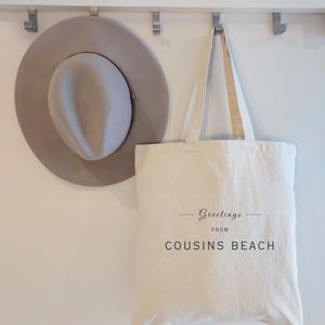 May include: A light gray fedora hat and a cream-colored tote bag hanging on a white wall. The tote bag has the words "Greetings from Cousins Beach" printed on it in black script.