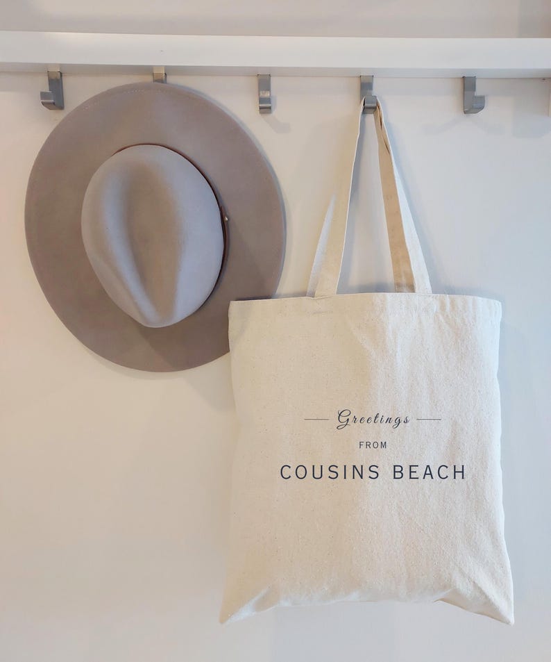 May include: A light gray fedora hat and a cream-colored tote bag hanging on a white wall. The tote bag has the words "Greetings from Cousins Beach" printed on it in black script.