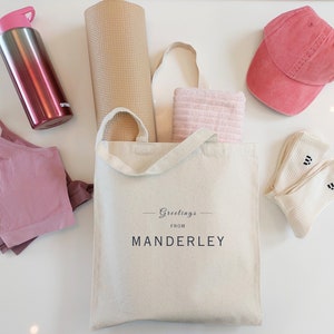 Beige canvas tote bag with the text Greetings from Manderley in black font. The bag is displayed on a white background and used as a gym bag filled with a yoga mat and towel, and is next to a sports bra, water bottle, cap and white socks.