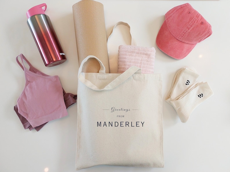 Beige canvas tote bag with the text Greetings from Manderley in black font. The bag is displayed on a white background and used as a gym bag filled with a yoga mat and towel, and is next to a sports bra, water bottle, cap and white socks.