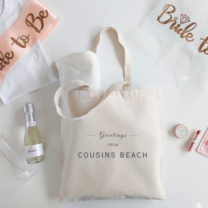 May include: A collection of bridal shower gifts, including a canvas tote bag with "Greetings from Cousins Beach" printed on it, a bottle of sparkling wine, and a "Bride to Be" sash.