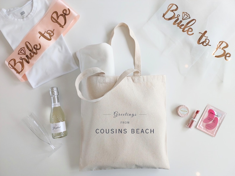 May include: A collection of bridal shower gifts, including a canvas tote bag with "Greetings from Cousins Beach" printed on it, a bottle of sparkling wine, and a "Bride to Be" sash.