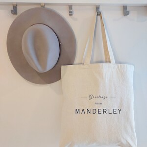 Beige canvas tote bag with the text Greetings from Manderley in black font.  The bag is hanging on a wall hook beside a stylish gray felt hat.