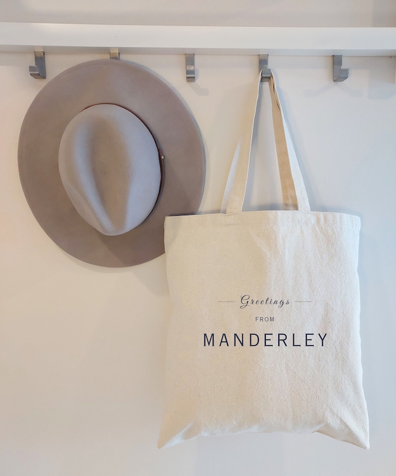 Beige canvas tote bag with the text Greetings from Manderley in black font.  The bag is hanging on a wall hook beside a stylish gray felt hat.