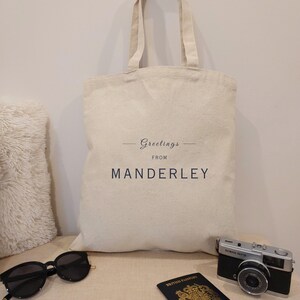 Beige canvas tote bag with the text Greetings from Manderley in black font. The bag is displayed against a neutral background with travel essentials including a passport, vintage camera and black sunglasses.