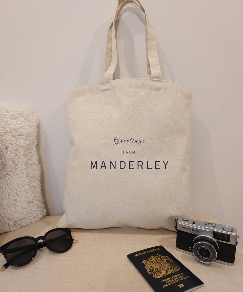 Beige canvas tote bag with the text Greetings from Manderley in black font. The bag is displayed against a neutral background with travel essentials including a passport, vintage camera and black sunglasses.