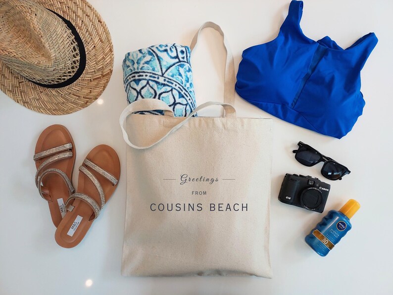 May include: A beach-themed flat lay featuring a straw hat, sandals, a tote bag with "Greetings from Cousins Beach" text, a blue swimsuit top, sunglasses, a camera, a sunscreen bottle, and a patterned towel.