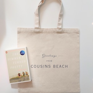 May include: A beige canvas tote bag with the text "Greetings from Cousins Beach" and a book titled "The Summer I Turned Pretty" by Jenny Han. The book cover features a beach scene with three people running.
