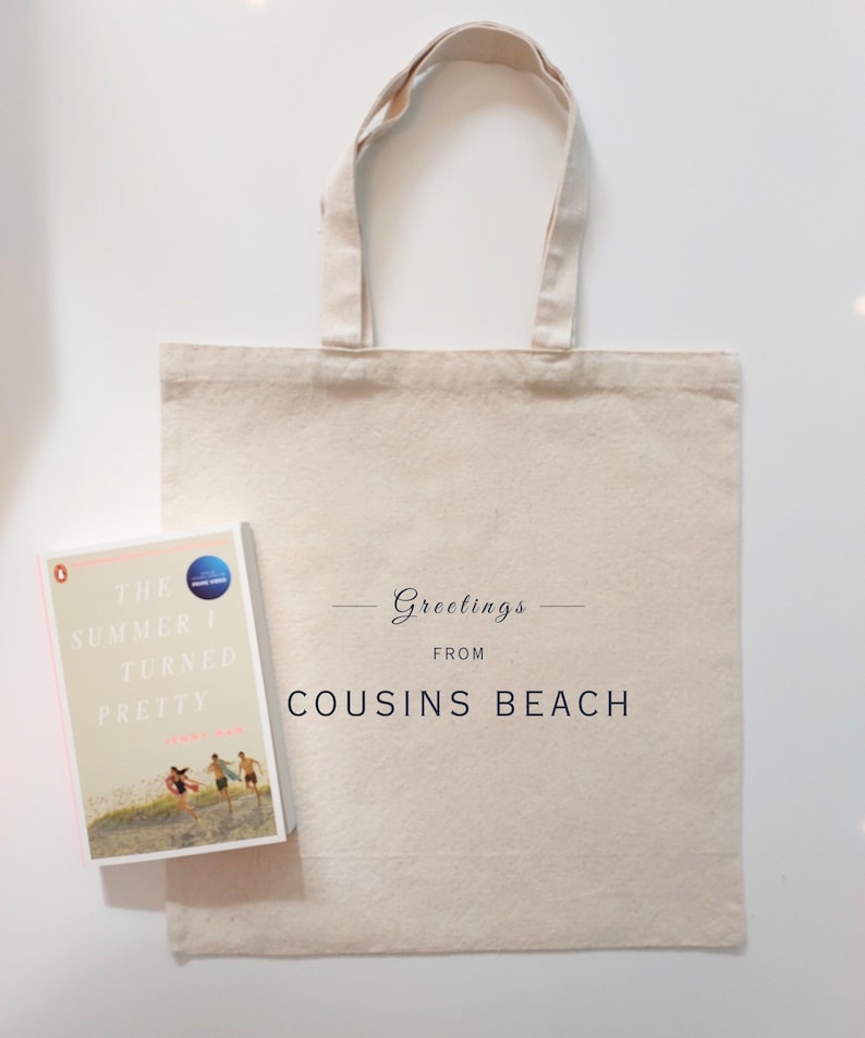 May include: A beige canvas tote bag with the text "Greetings from Cousins Beach" and a book titled "The Summer I Turned Pretty" by Jenny Han. The book cover features a beach scene with three people running.