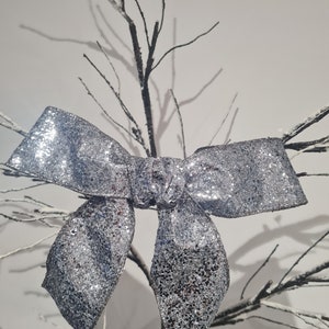 May include: A silver glitter bow with a large centre loop and two smaller loops on either side. The bow is tied to a branch of a white-painted tree.