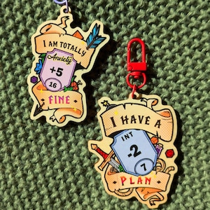 Puede incluir: Dos llaveros de madera con un diseño de estilo de dibujos animados. El primer llavero presenta un rollo con el texto "I am totally anxiety +5 16 FINE". El segundo llavero presenta un rollo con el texto "I have a INT -2 1 PLAN".