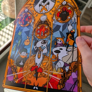 May include: A stained-glass style art print featuring characters from the video game Hollow Knight. The artwork has a gold border and depicts various characters in a colourful, geometric design. The print has the text "Cattering Art" at the bottom.