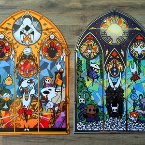 May include: Two stained-glass style art prints depicting video game characters. One has an orange border and warm hues, the other a dark blue border and cool tones. Both feature detailed designs and geometric patterns, reminiscent of church windows.